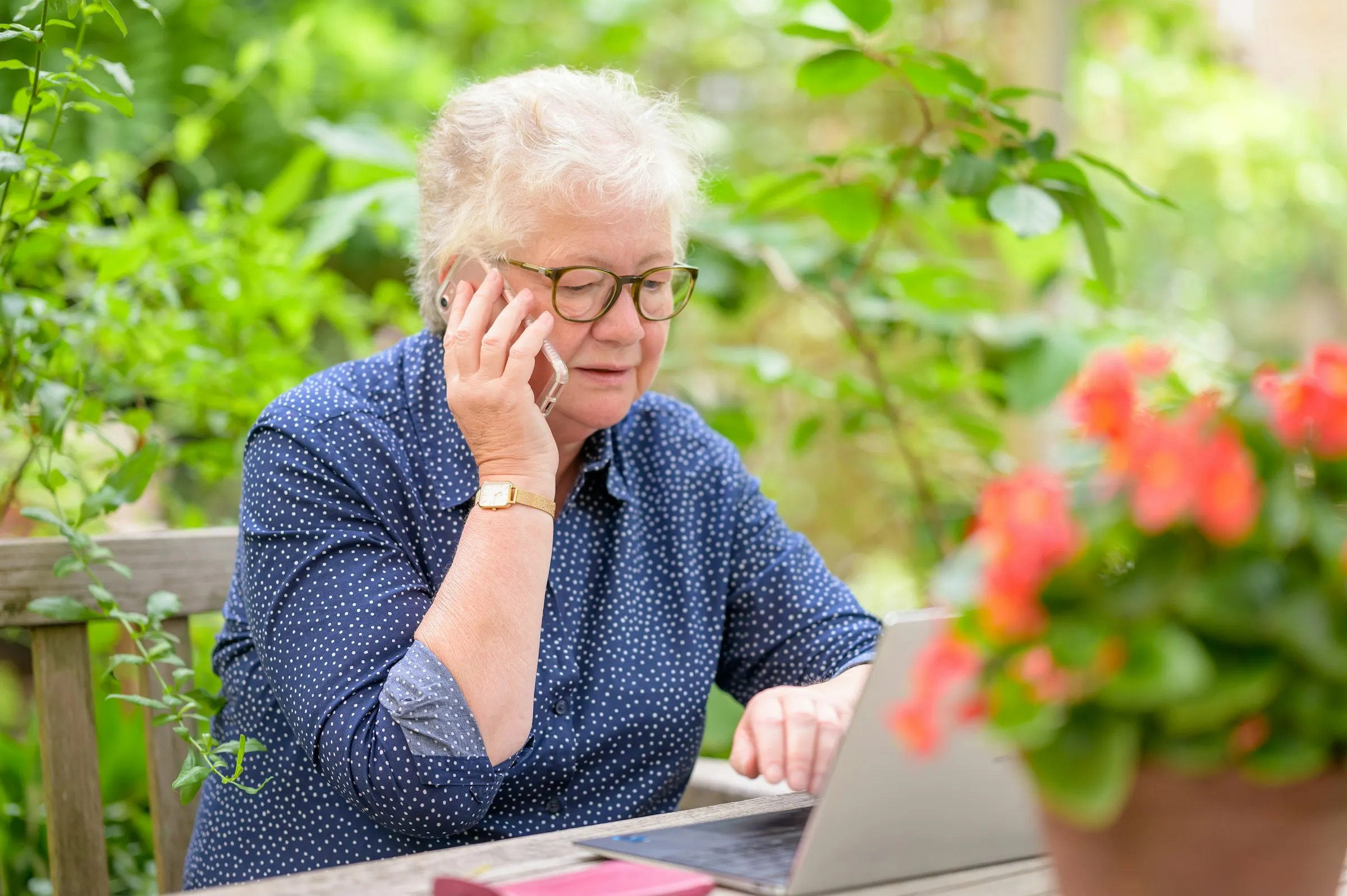 An older woman on the phone reviewing investments on her laptop at a table in a sunny garden