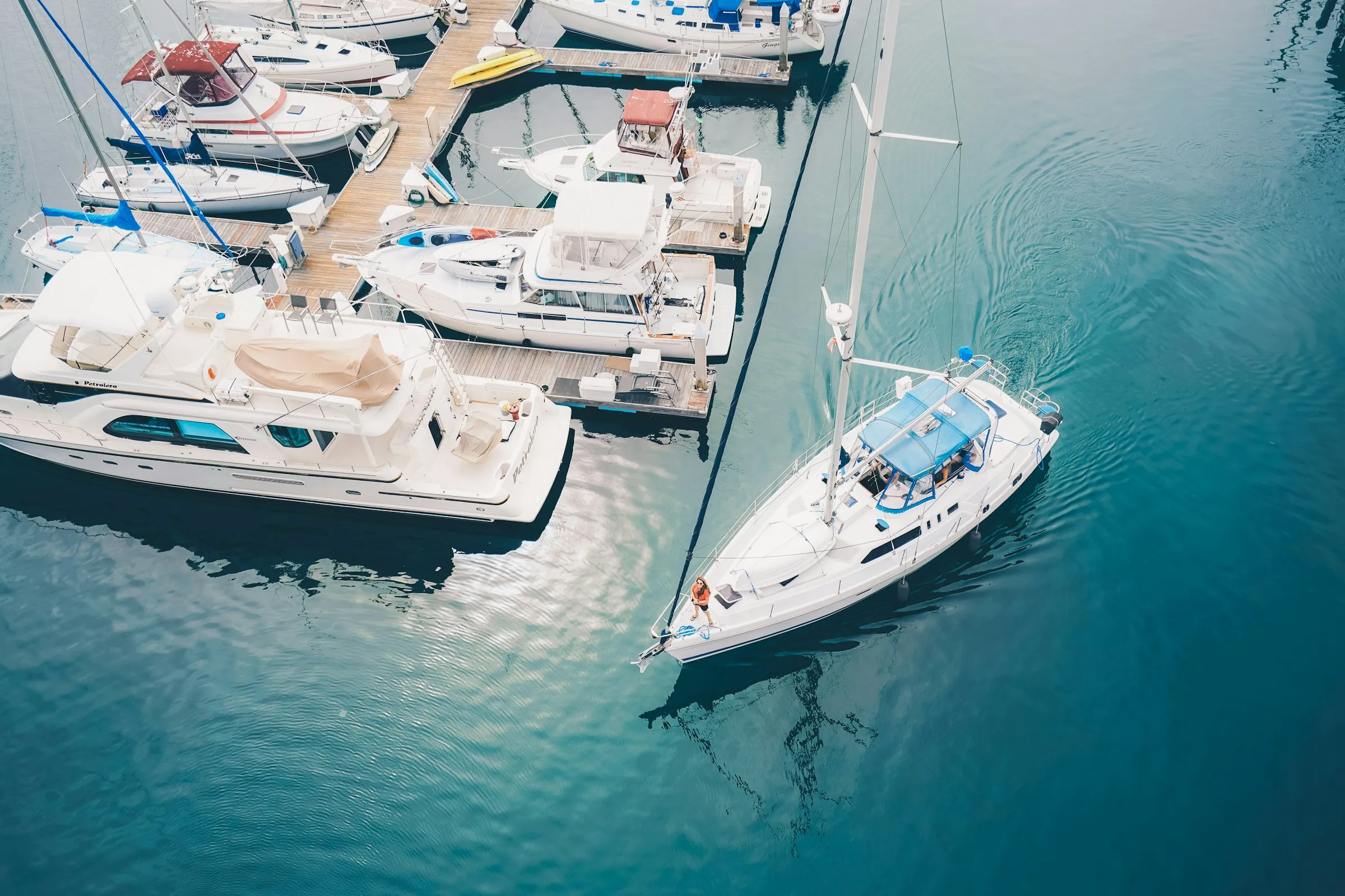 Aerial view of launches and yachts berthed in a sunny marina, with a yacht moving through clear blue-green water
