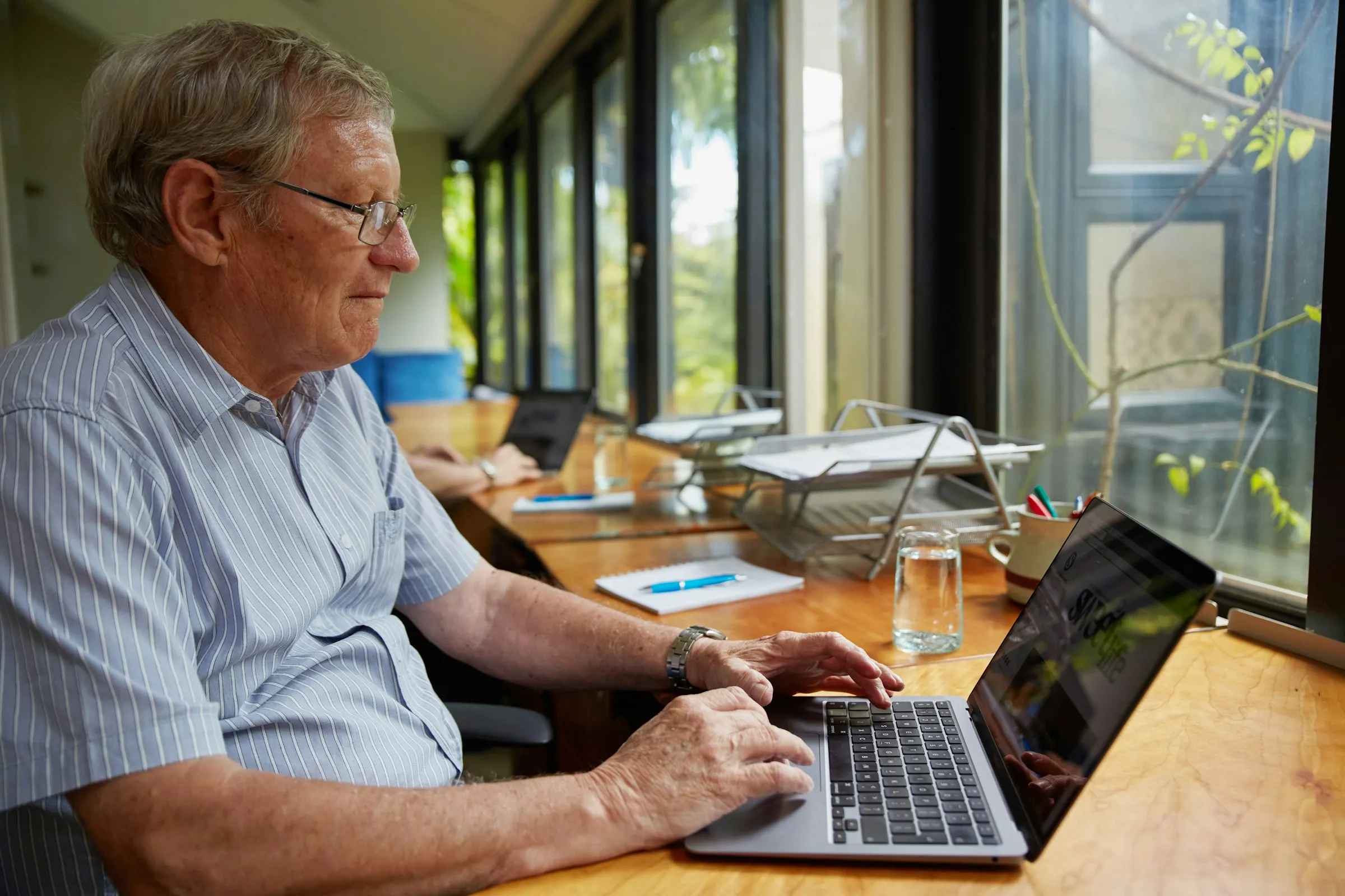 A retired man in glasses reviewing a loan pack on his laptop at a sunny window desk